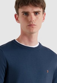 Granatowy dzianinowy sweter z prążkowaną okrągłą szyją i dyskretnym pomarańczowym logo na klatce piersiowej; gładka faktura i dopasowane rękawy.