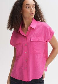 Camicetta rosa a maniche corte con texture sgualcita, apertura frontale con bottoni e due tasche sul petto. Vestibilità casual con orlo leggermente curvo.