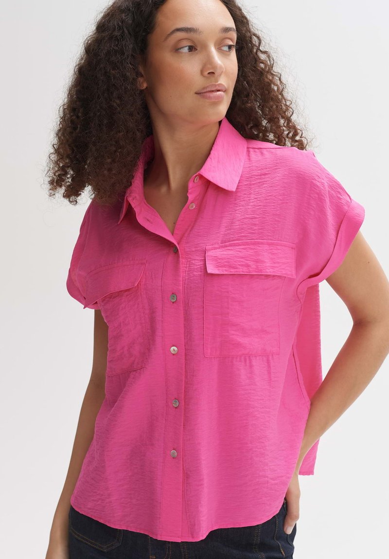 Camicetta rosa a maniche corte con texture sgualcita, apertura frontale con bottoni e due tasche sul petto. Vestibilità casual con orlo leggermente curvo.