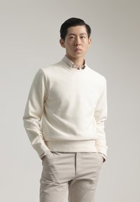 Sudadera de color crema claro con textura acanalada y cuello redondo, que presenta un logo sutil. Combinada con pantalones beige claros.
