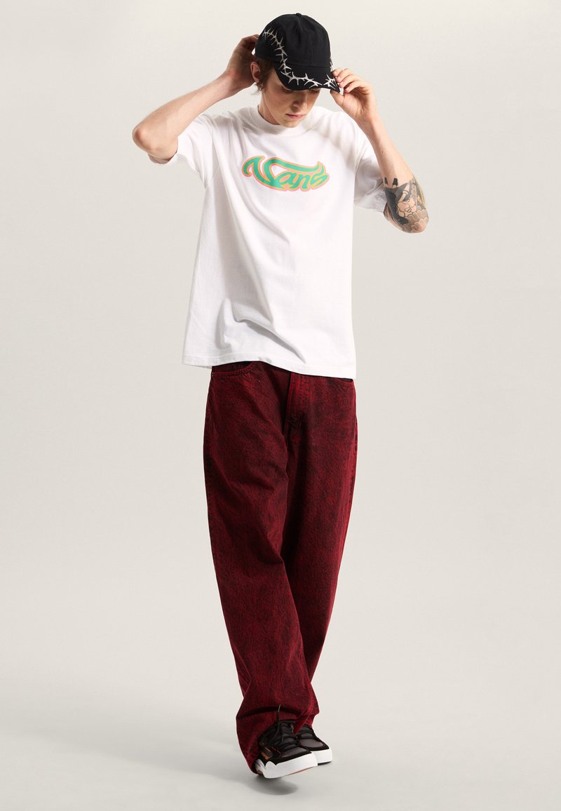 T-shirt blanc avec logo "Vans" multicolore, associé à un pantalon rouge ample avec un motif texturé, et des baskets noires avec des semelles blanches.