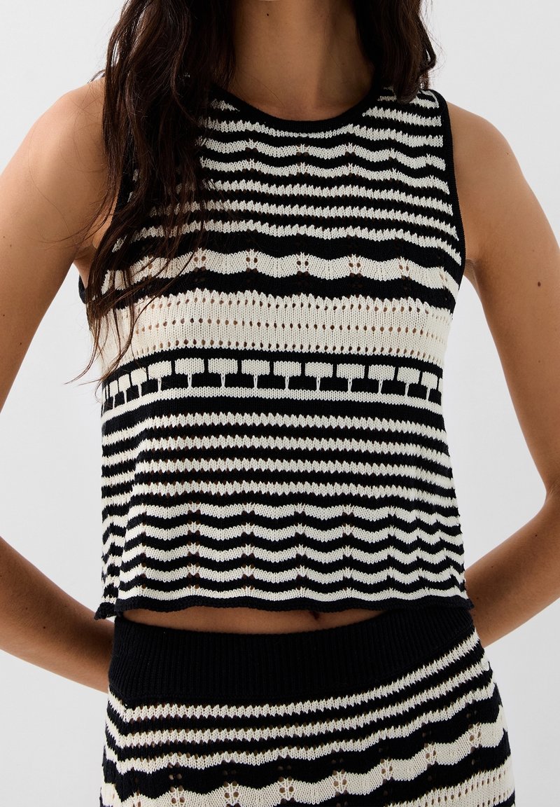 Femme portant un haut court sans manches en maille noir et blanc avec une jupe taille haute assortie, présentant des motifs rayés et géométriques.