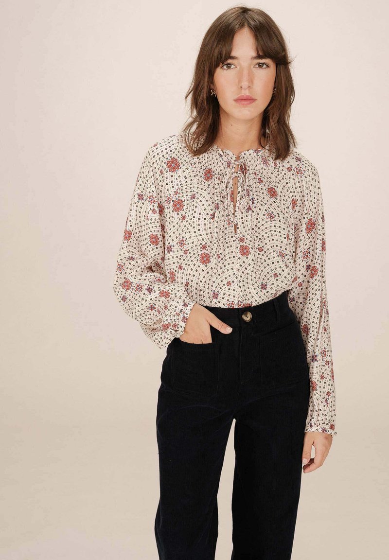 Blouse à imprimé floral en crème avec un col à nouer et de longues manches bouffantes, associée à un pantalon en velours côtelé noir taille haute.