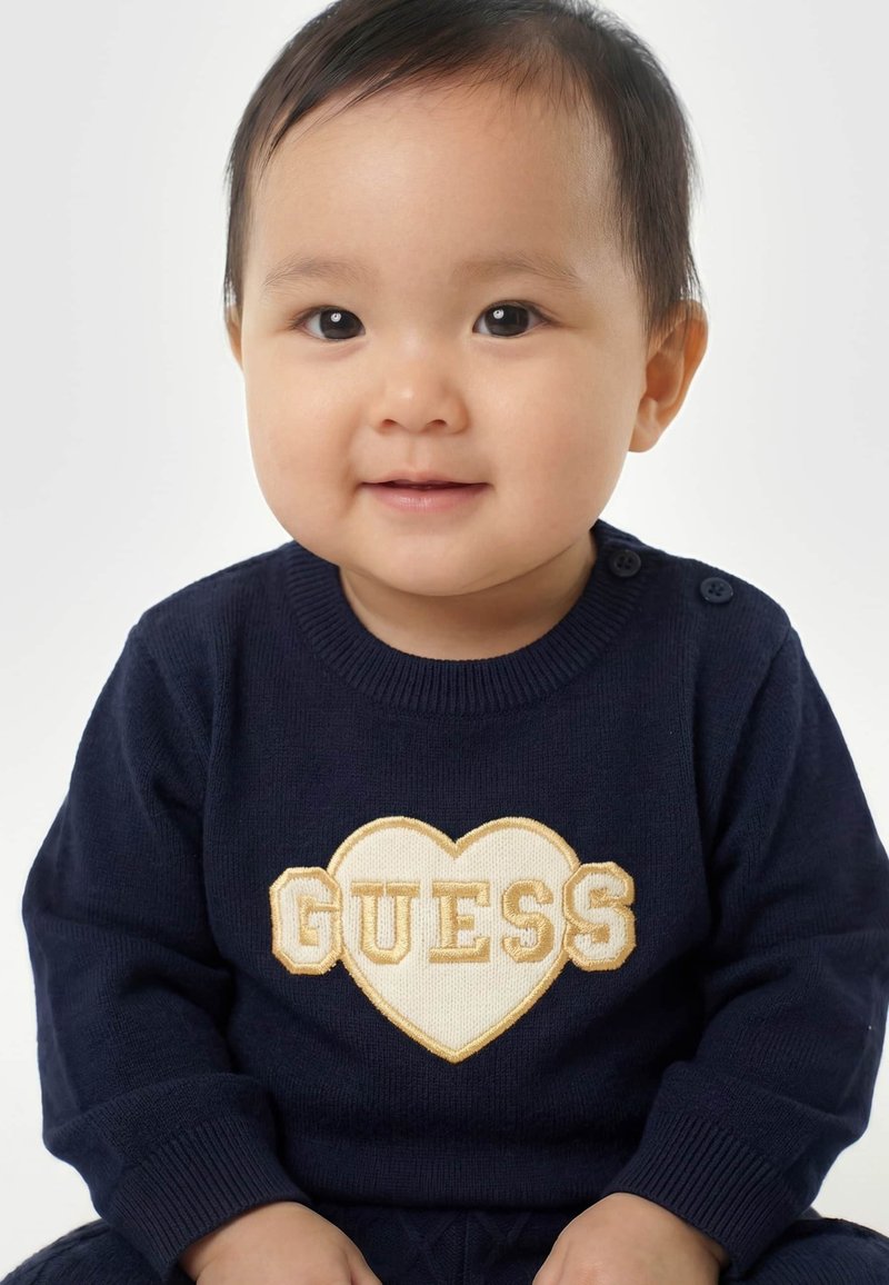 Tout-petit souriant aux cheveux courts et foncés, portant un pull bleu marine avec un cœur crème et le mot "GUESS" sur le devant, sur fond blanc.