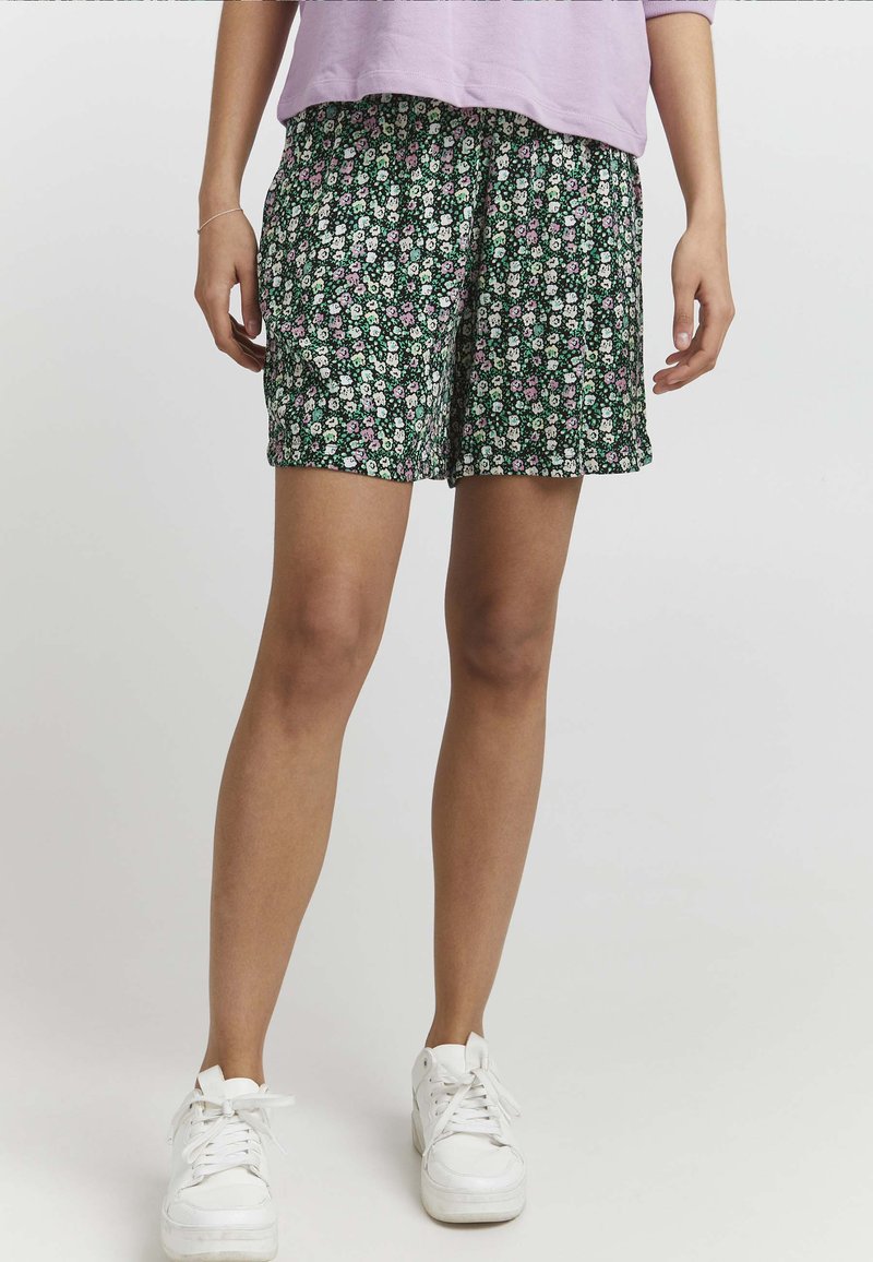 Shorts à imprimé floral vert foncé avec des fleurs blanches et roses. Tissu léger, coupe ample, avec un waistband élastiqué. Associés à des baskets blanches.