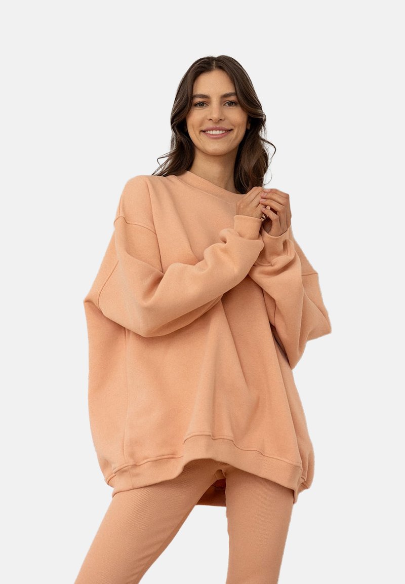 Przesadnie duża brzoskwiniowa bluza z okrągłym dekoltem, przesuniętymi ramionami i miękką teksturą, w połączeniu z dopasowanymi, obcisłymi spodniami.
