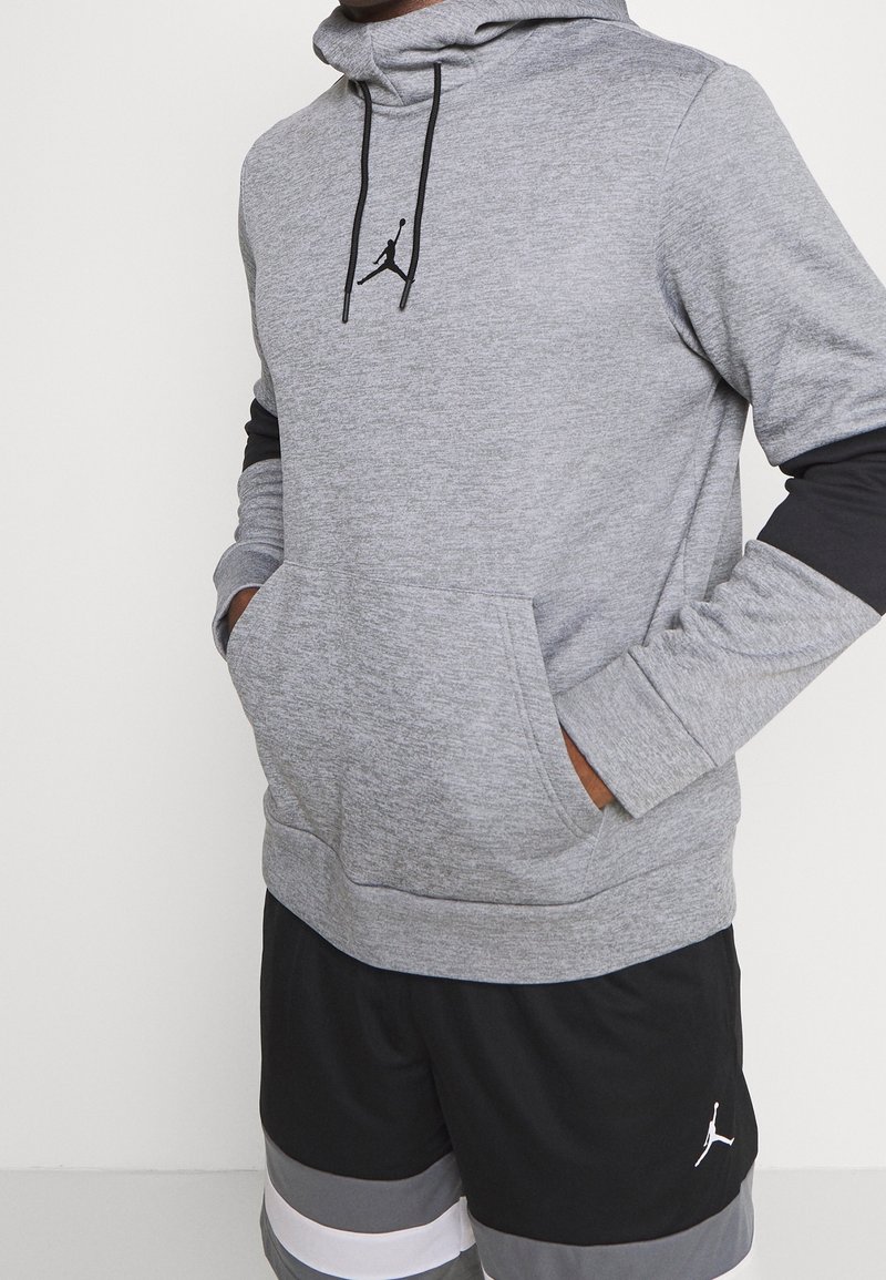 Šedá mikina s kapucí vyrobená z měkké, strukturované tkaniny. Má přední kapsu, černé detaily a černé logo Jumpman na hrudi.
