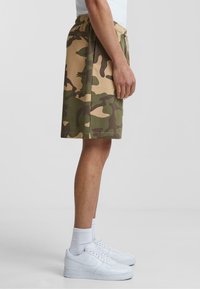 Camo-print shorts in shades of green and brown, with an elastic waistband. Worn with white sneakers and ankle socks.