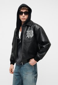 Hombre con sudadera negra con capucha bajo chaqueta de cuero negra con bordado gótico blanco, vaqueros azules y gafas de sol negras.