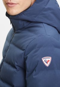 Námořnicky modrá péřová bunda s kapucí, s výrazným logem Rossignol na rameni, hladkou texturou a prošívaným designem.