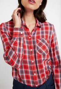 Chemise à carreaux rouge et noir à boutons, avec une poche poitrine, des boutons blancs et des manches longues en tissu texturé.