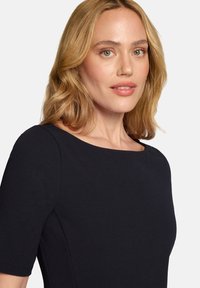 Femme blonde aux yeux vert clair portant un haut noir à manches courtes, regardant légèrement sur le côté avec une expression faciale neutre.