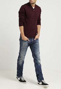 Camisa henley de manga larga color burdeos con cuello abotonado, combinada con jeans azules desgastados y zapatillas en blanco y negro.