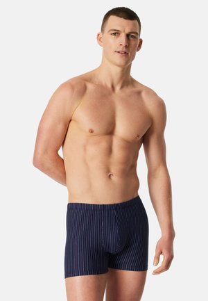 Uomo muscoloso in piedi, che indossa boxer blu navy con sottili strisce verticali, posando contro uno sfondo chiaro e uniforme.