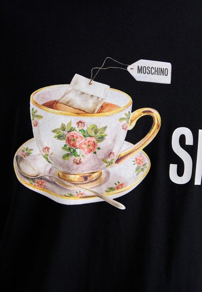 T-shirt graphique avec une tasse à thé ornée de motifs floraux dans des couleurs pastel, un sachet de thé et une cuillère, imprimée sur un fond en tissu noir.