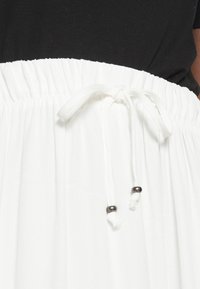 Jupe blanche avec taille élastiquée froncée et cordon noué avec deux perles argentées, portée avec un haut noir.
