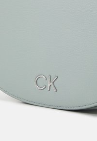 Borsa a spalla in pelle azzurro chiaro con logo "CK" in argento, texture liscia, forma tondeggiante e dettagli di cucitura a contrasto.