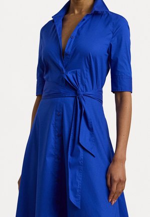 Kvinde iført en knaldblå knap-op kjole med albuelange ærmer og et bindebælte i taljen foran en ensfarvet baggrund.