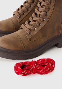 Bottines en cuir marron à lacets avec des semelles épaisses, posées à côté d'un lacet rouge et blanc enroulé sur une surface blanche.