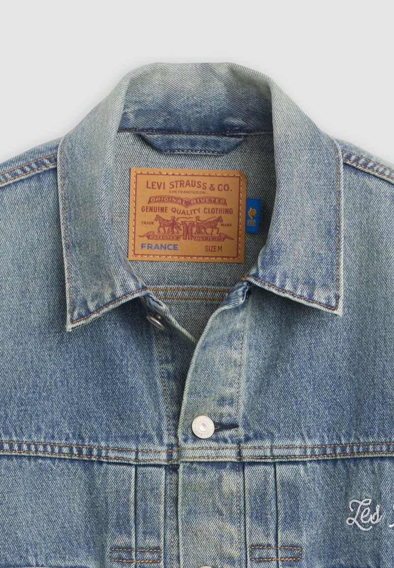 Collo della giacca in denim con etichetta in pelle Levi Strauss & Co. visibile, etichetta blu, bottoni in metallo e testo ricamato su tessuto lavato azzurro chiaro.