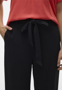 Pantaloni neri a vita alta con cintura da annodare, dotati di tasche laterali e una texture del tessuto liscia. Abbinati a una maglietta rossa a maniche corte.