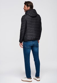 Chaqueta negra acolchada con capucha, con textura de acolchado horizontal. Combinada con jeans azules y zapatillas blancas. Fondo neutro.