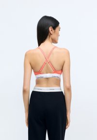 Korallszínű bralette keresztpántos dizájnnal, fehér Calvin Klein logóval ellátott övvel. A fekete nadrág kontrasztos derekával egészíti ki a megjelenést.