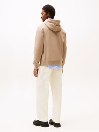 Felpa marrone chiaro con tasca posteriore, indossata sopra una maglietta a righe blu e bianche. Abbinata a pantaloni a gamba larga color crema e scarpe nere.