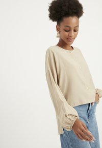 Beige verkürztes Shirt mit langen, gepufften Ärmeln und Knopfleiste vorne. Der Stoff wirkt weich und hat eine glatte Textur.