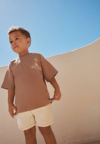 Un niño pequeño con una camiseta marrón de gran tamaño y pantalones cortos blancos está de pie al aire libre frente a una pared curva beige y un cielo azul claro.