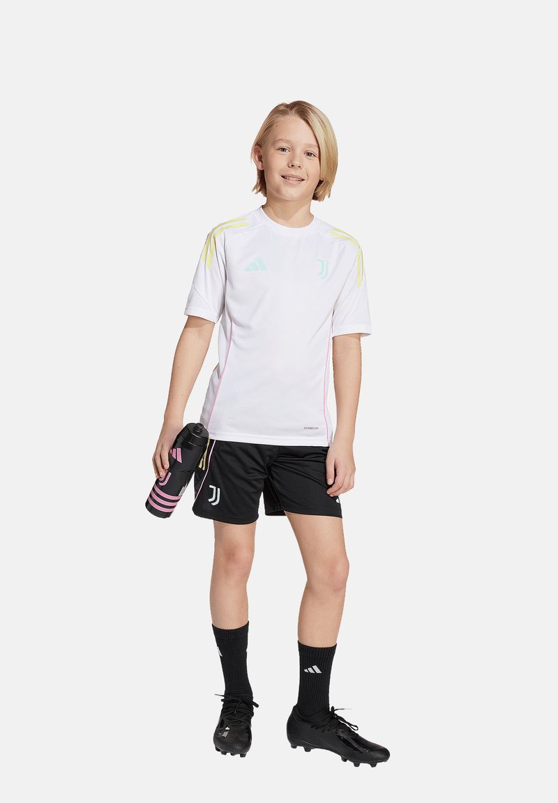 Jugendlicher in einem weißen Sporttrikot mit gelben Akzenten und schwarzen Shorts. Hält eine schwarze Wasserflasche mit pinken Streifen. Fußballschuhe sind sichtbar.