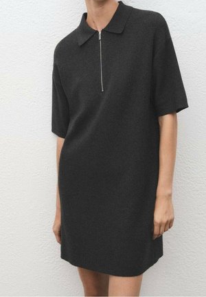 Personne portant une robe polo noire à manches courtes avec une fermeture éclair sur le devant, debout contre un mur clair texturé.