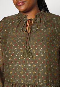 Olijfgroene blouse met een ruches halslijn, een franjen strikdetail en kleine bloemenpatronen in het rood, geel en blauw door de stof heen.