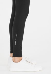 Mustat legginsit, joissa on sileä pinta, ja vasemmalla puolella valkoinen teksti "UUSI HUONOT TOIMINNOT". Asusteena valkoiset lenkkarit.