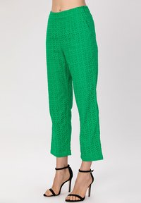 Pantalons légers verts avec un design perforé, présentant une coupe droite et une taille mi-haute. Associés à des sandales à talons hauts noires.
