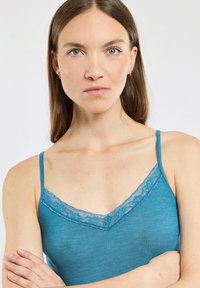 Jeune femme aux cheveux bruns raides portant un débardeur en dentelle turquoise, bras croisés, regardant directement l'appareil photo avec une expression neutre.