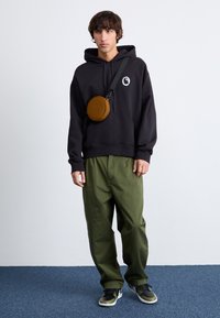 Sudadera negra con un logo, pantalones verde oliva y zapatillas. Se lleva una mochila tipo bandolera marrón cruzada sobre el cuerpo. Fondo liso.