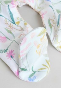 Pehmed valged beebikindad pastellsete lillemustriste ja sisselõigetega, avatud asendis heleda halli pinna peal.