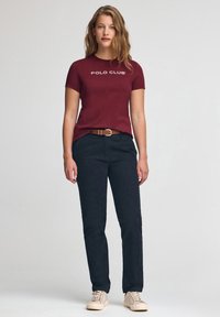 Camiseta de manga corta color granate con el logo "POLO CLUB", combinada con pantalones azul marino oscuro y zapatillas beige, acentuada con un cinturón marrón.