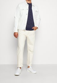 Giacca di denim bianca, maglione navy con pois, pantaloni beige e sneakers bianche. Le caratteristiche principali includono chiusura con bottoni, pantaloni con risvolto e design casual.