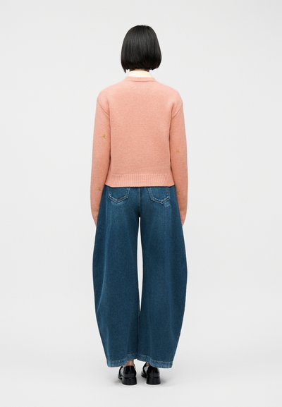 Άτομο με κοντά μαύρα μαλλιά, φορώντας ροζ πουλόβερ, φαρδιά μπλε τζιν και μαύρα παπούτσια, στέκεται γυρισμένο με την πλάτη σε απλό φόντο.