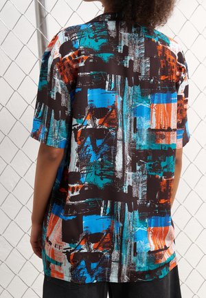 Personne portant une chemise à manches courtes avec un motif abstrait en bleu, orange, noir et blanc, debout devant une clôture en grillage.