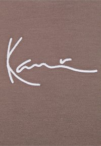 Brązowy materiał o gładkiej fakturze, z białym haftowanym podpisem. Wzór jest minimalistyczny i umieszczony centralnie na tkaninie.