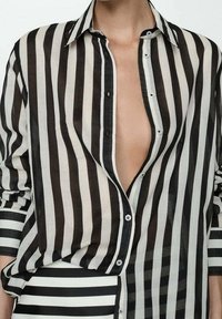 Camicia a righe bianco e nero con una vestibilità rilassata, tessuto trasparente e scollatura a V profonda; presenta righe orizzontali e verticali contrastanti.