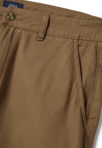 Pantalones chinos color arena mostrando la cintura con botón, pasador de cinturón, bolsillo lateral y una etiqueta azul que dice "CHINOS" en el interior.