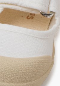 Zapato de lona blanco con suela de goma beige, que presenta una correa con un acento metálico plateado y un detalle de tejido texturizado en la zona de los dedos.