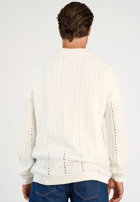 Man draagt een witte, gebreide trui met lange mouwen en een verticale textuur, gezien van achteren, gecombineerd met een blauwe spijkerbroek.