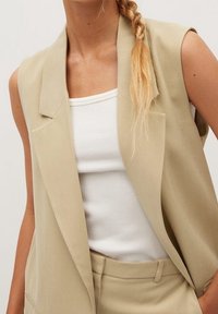 Beżowa bezrękawnikowa marynarka z klapami, noszona na białym topie. Gładki materiał o dopasowanym kroju, bez widocznych wzorów czy ozdób.