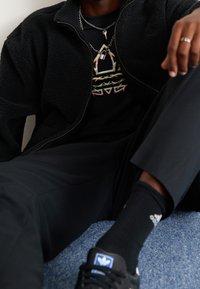 Chaqueta de forro polar negro con cremallera sobre una camiseta gráfica negra con patrones coloridos, combinada con pantalones negros, calcetines y zapatillas deportivas negras.