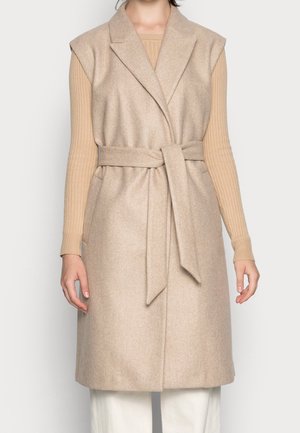 Beige mouwloze wollen vest met brede revers en zelfbindceintuur, gedragen over een lange mouw ribgebreide beige top en witte broek.